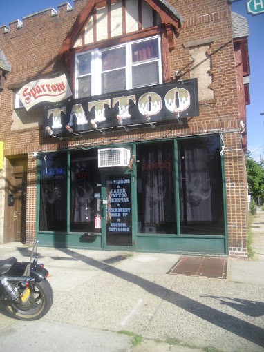 Street view of a tattoo parlor with a brick facade. A large black sign reading "Tattoo" in white letters adorns the entrance, hinting at its reputation for skilled tattoo artists in Nassau County NY. A motorcycle is parked on the sidewalk, next to a smaller sign advertising various tattoo services.