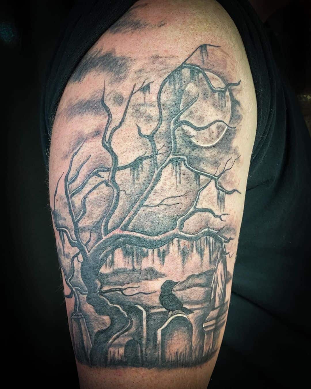 A tattoo on an upper arm, expertly crafted by a skilled tattoo artist in Nassau County NY, features a detailed, eerie landscape with a leafless tree, rocky formations, and a silhouette of a perched raven. The cloudy sky background completes the mysterious gothic atmosphere.