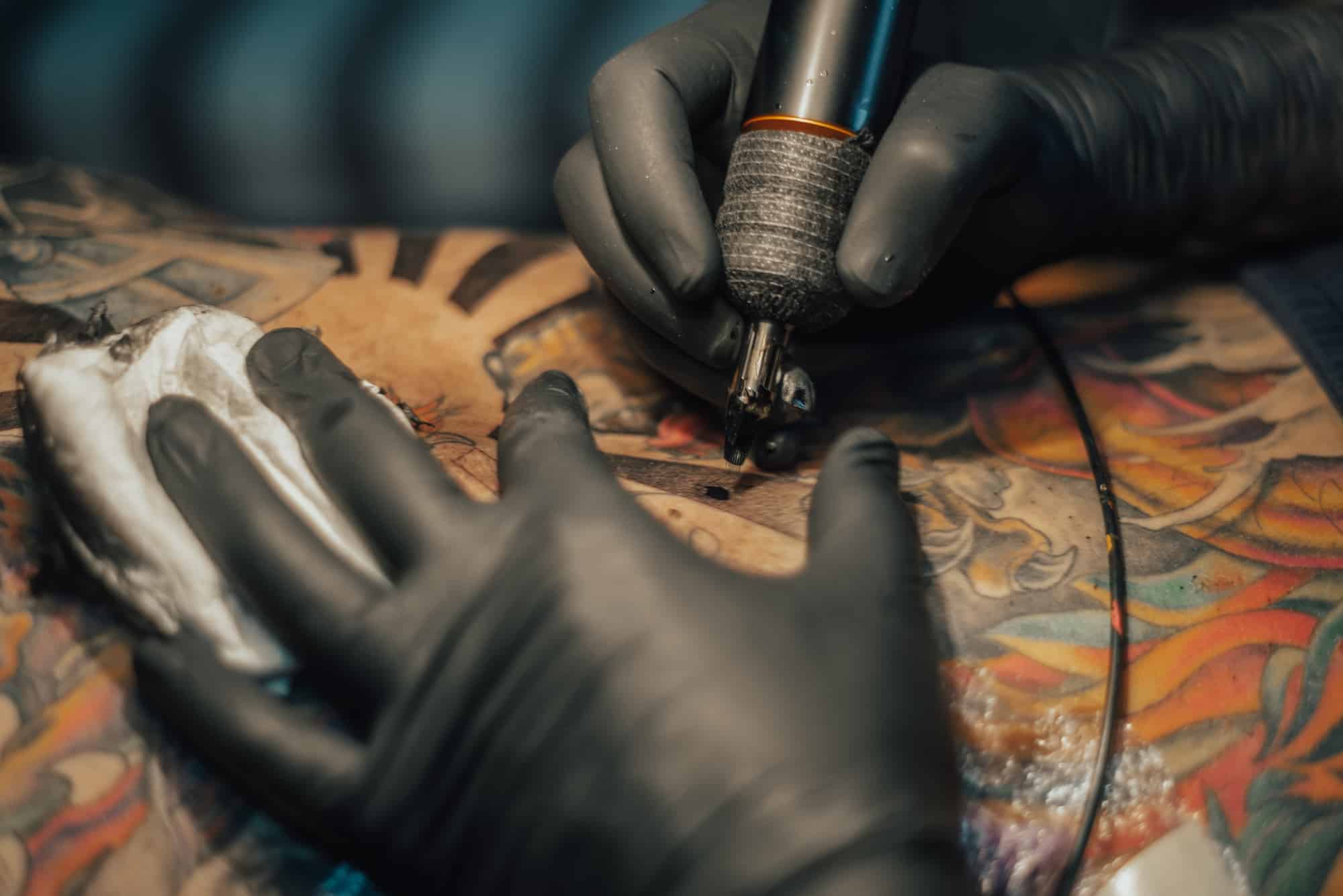 A tattoo artist in Nassau County, NY, wearing black gloves, meticulously uses a tattoo machine to add intricate details to a colorful design on a person’s skin. A paper towel is used to wipe away excess ink nearby.
