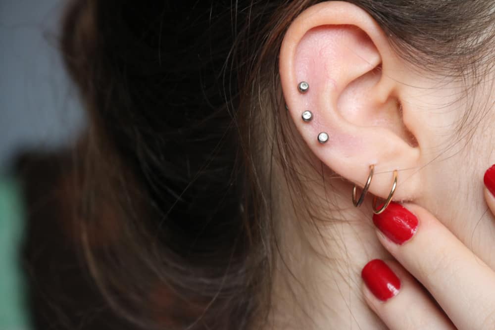 Close-up of a person's ear with three small stud piercings along the cartilage from a piercing shop in Nassau County NY. They are wearing two gold hoop earrings in the lower lobe. The person has red nail polish and is touching their ear.