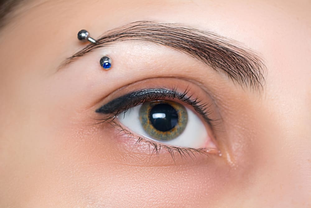 Close-up of a person’s eye with bold eyeliner and a blue-eyed eyebrow piercing above it. The curved metallic bar, featuring a small blue gemstone, showcases the expertise found at piercing shops in Nassau County, NY. The skin is smooth, and the eyelashes are naturally curled.
