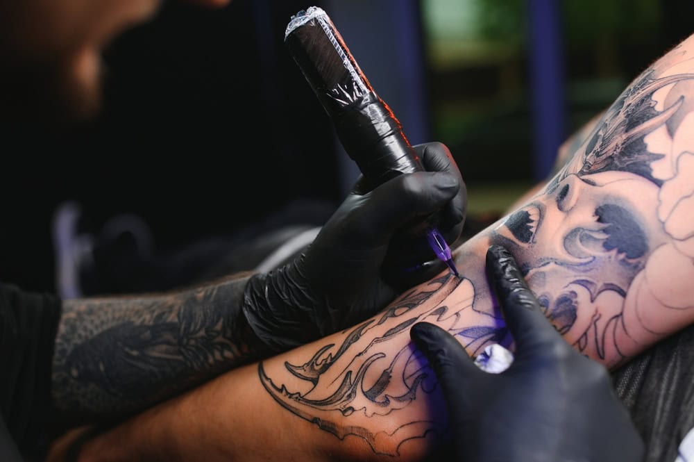 A tattoo artist with gloves is using a tattoo machine to ink a detailed design on a person's arm. The design features intricate patterns, and the scene is captured in close-up. The artist's arm also shows existing tattoos.